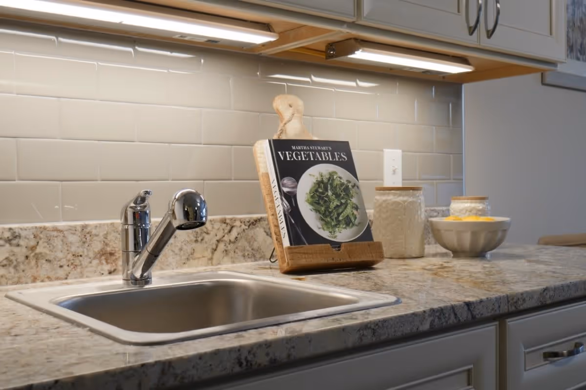 A kitchen countertop with a stainless steel sink and a modern faucet. On the countertop, there is a cookbook titled 'Martha Stewart's Vegetables' displayed on a wooden stand, along with two decorative jars and a bowl. The backsplash features light-colored subway tiles, and there are under-cabinet lights illuminating the area.