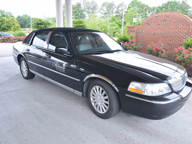 A black sedan with a 'Rosewood' logo parked under an entrance portico in front of a brick landscaped area.