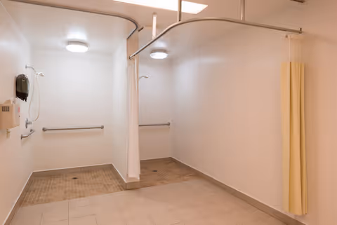 Clinical shower room with two roll-in shower stalls, curtain dividers, grab bars, and tiled floors.