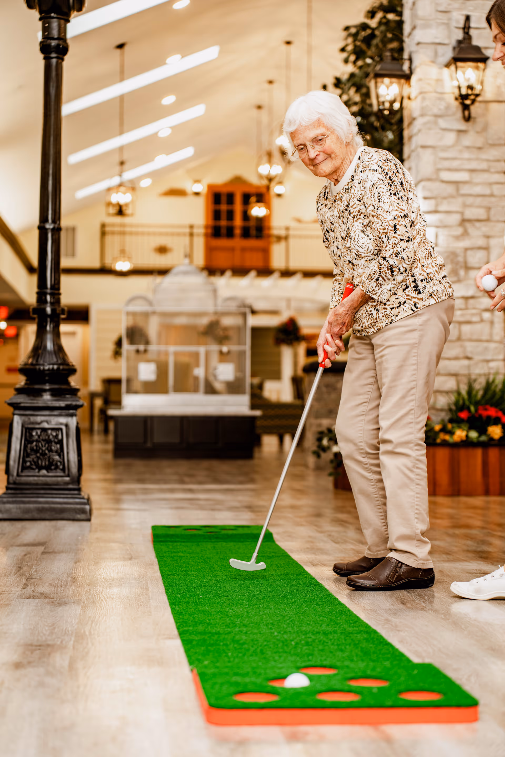 An elderly woman playing indoor mini golf on a green putting mat inside a well-lit assisted living facility with a high ceiling, decorative lamps, and stone walls. She is holding a putter and aiming at a golf ball near several holes on the mat.