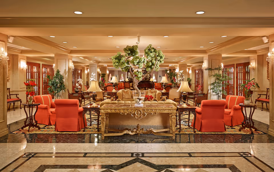 Ornate, well-lit lobby/lounge with red upholstered armchairs, sofas, side tables, lamps, and decorative plants arranged around a central ornate table.