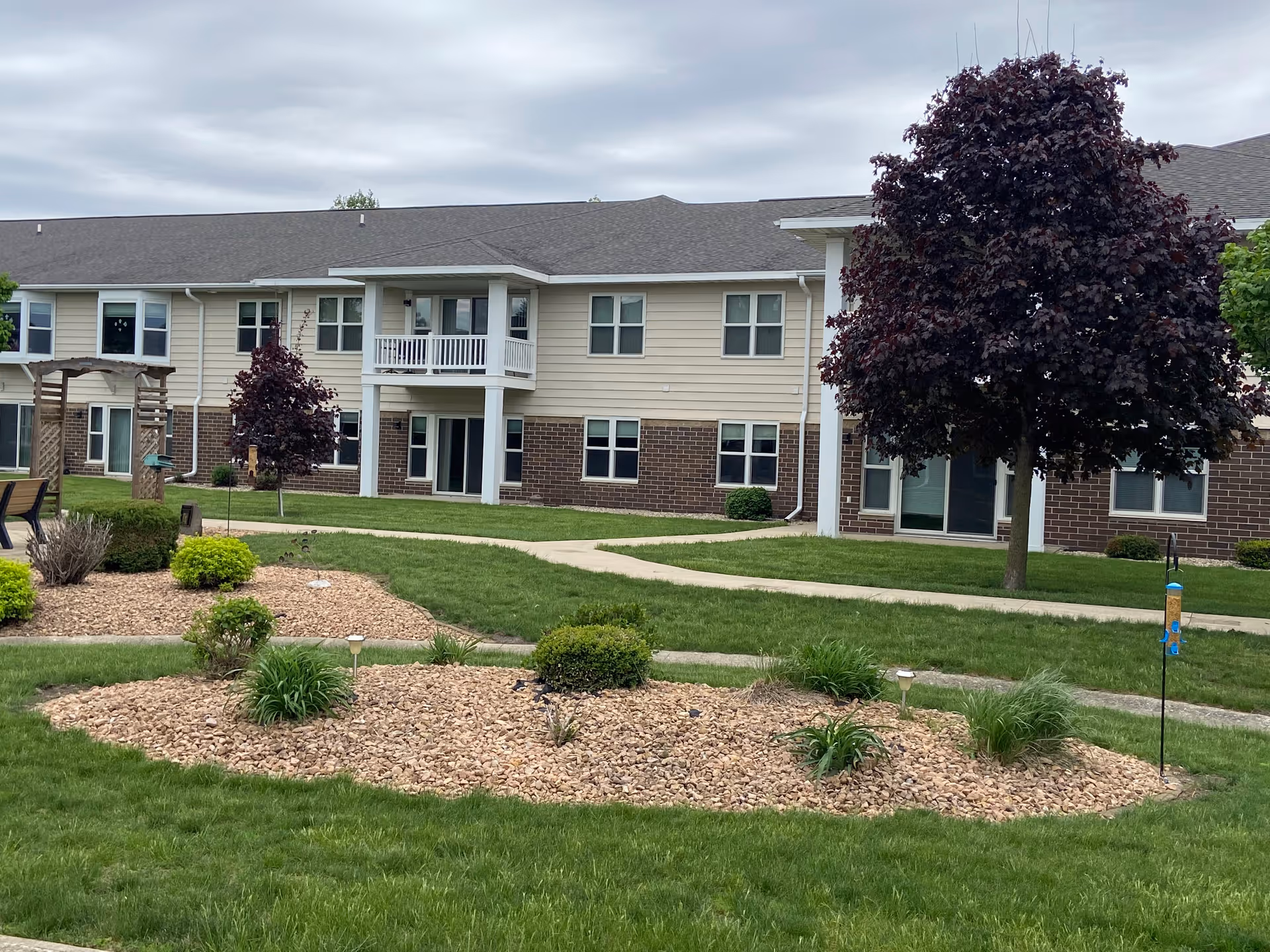 Outdoor view of The Meadows Senior Community showing a two-story building with beige siding and brick accents. The foreground features a well-maintained lawn with landscaped garden beds containing small bushes and plants, a paved walkway, and a tree with dark red leaves. The sky is overcast.