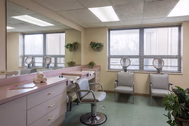 Interior view of a salon area in a care facility with two salon chairs in front of a long counter with sinks and mirrors. Two additional chairs are placed near a window with blinds. The room has light-colored walls, green flooring, and some potted plants on the walls and floor.