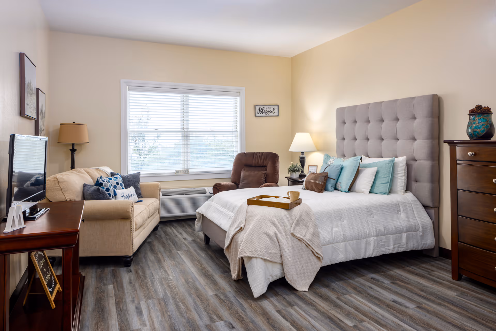 A cozy bedroom in a senior living facility featuring a large bed with a tufted headboard, multiple pillows, and a beige blanket. Next to the bed is a wooden nightstand with a lamp and decorative items. Across from the bed is a beige sofa with decorative pillows, a brown recliner chair, and a flat-screen TV on a wooden stand. The room has wood-look flooring, a large window with blinds, and light-colored walls with framed artwork.