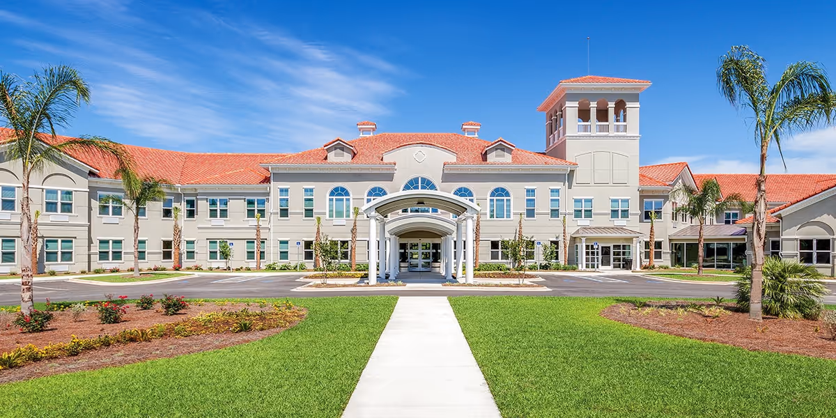 Front exterior view of Somerby Santa Rosa Beach facility with a wide concrete walkway leading to the main entrance, surrounded by green grass, landscaped flower beds, and palm trees under a blue sky.