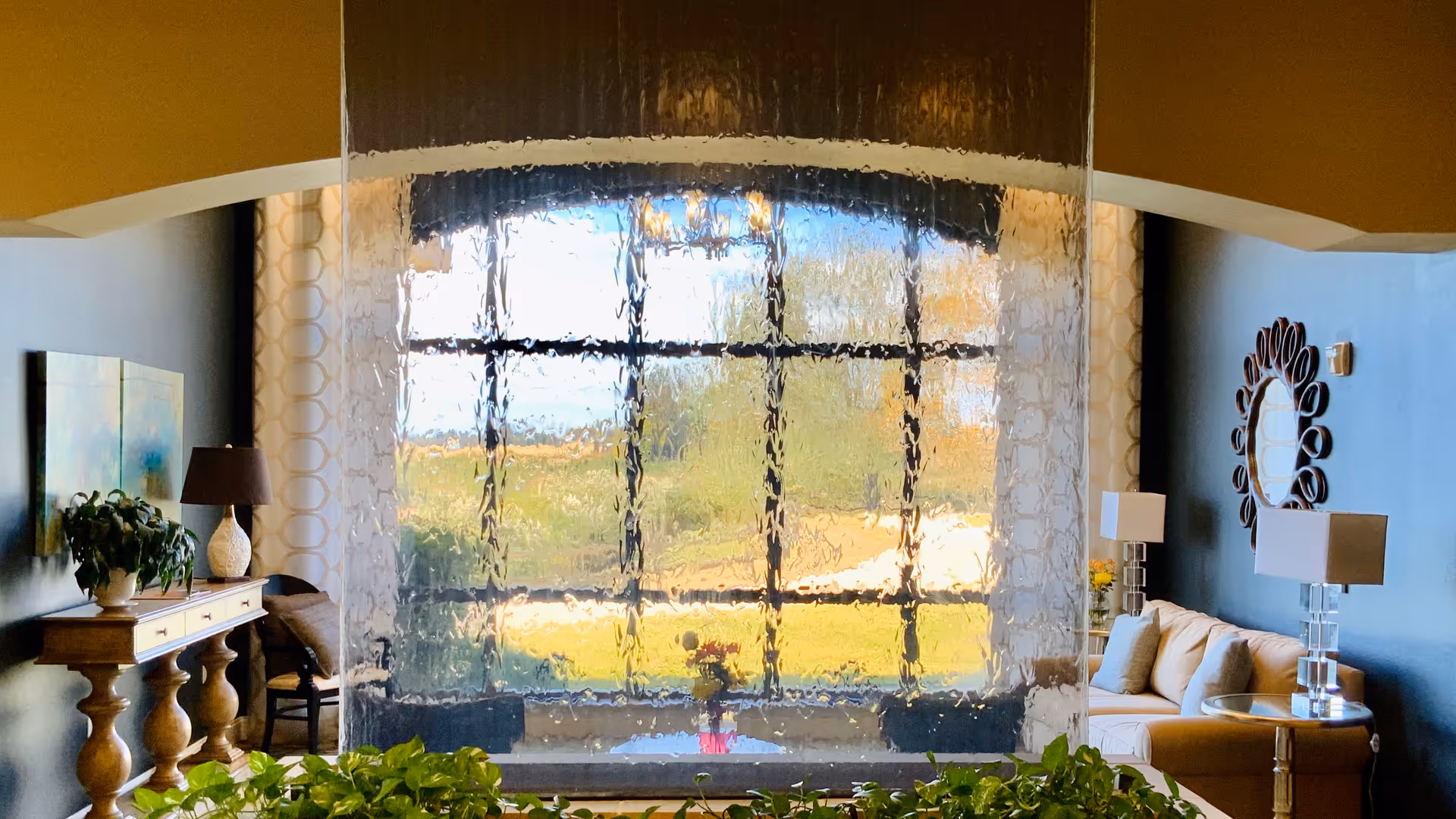 A bright living room lounge featuring a central glass water wall in front of a large window, flanked by sofas, lamps, plants, and decorative accents.