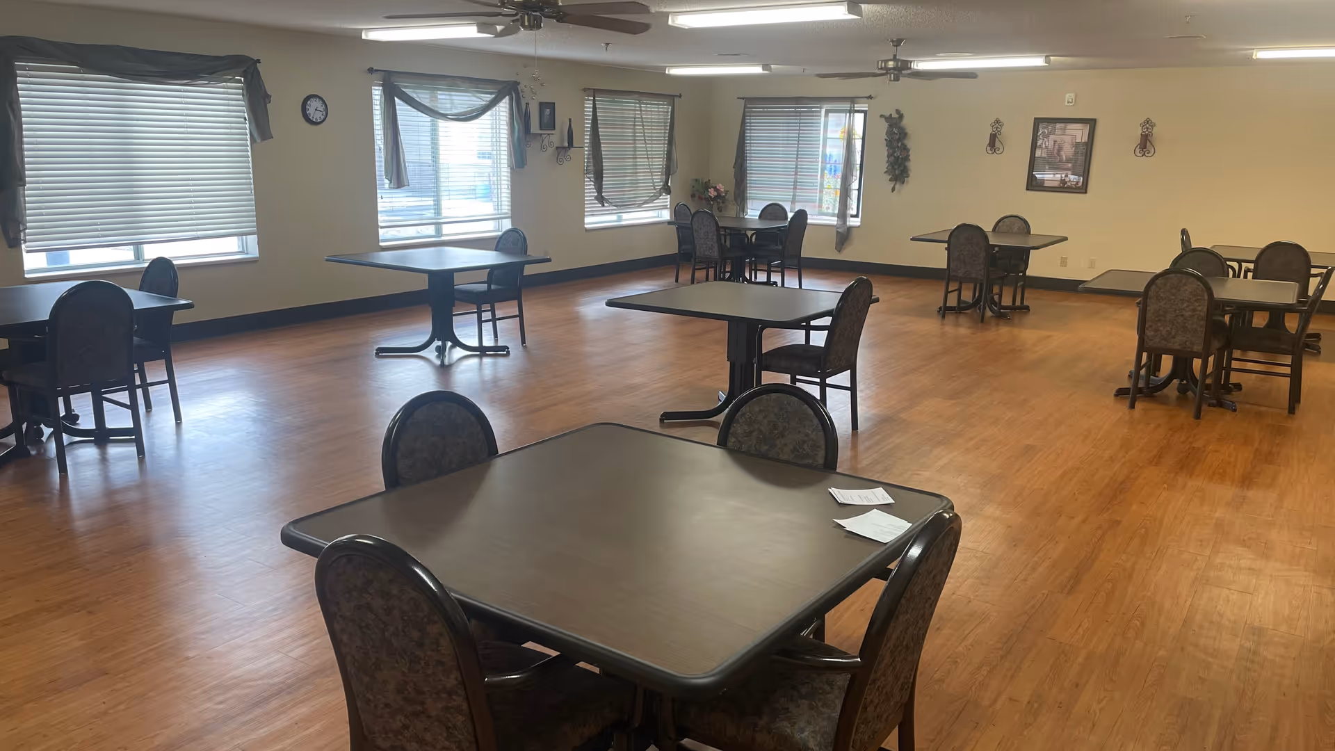Spacious common dining/activity room with multiple square tables and chairs on wood-look flooring and windows along the far wall.