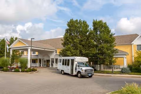 Carriage Green at Milford exterior: yellow building with covered entry, shuttle bus, and trees.