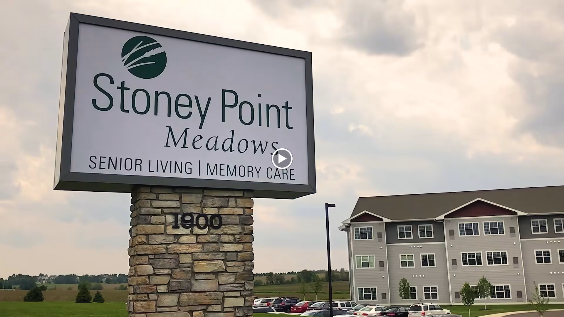 Large Stoney Point Meadows senior living sign on a stone pillar in front of a multi-story building under a cloudy sky.