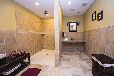 A spacious bathroom with beige tiled walls and floor. The room features a walk-in shower with a black showerhead, a bench with folded towels, a granite countertop with a sink and faucet, a mirror above the sink, and a laundry basket. The walls have two framed pictures, and the lighting is recessed in the ceiling.
