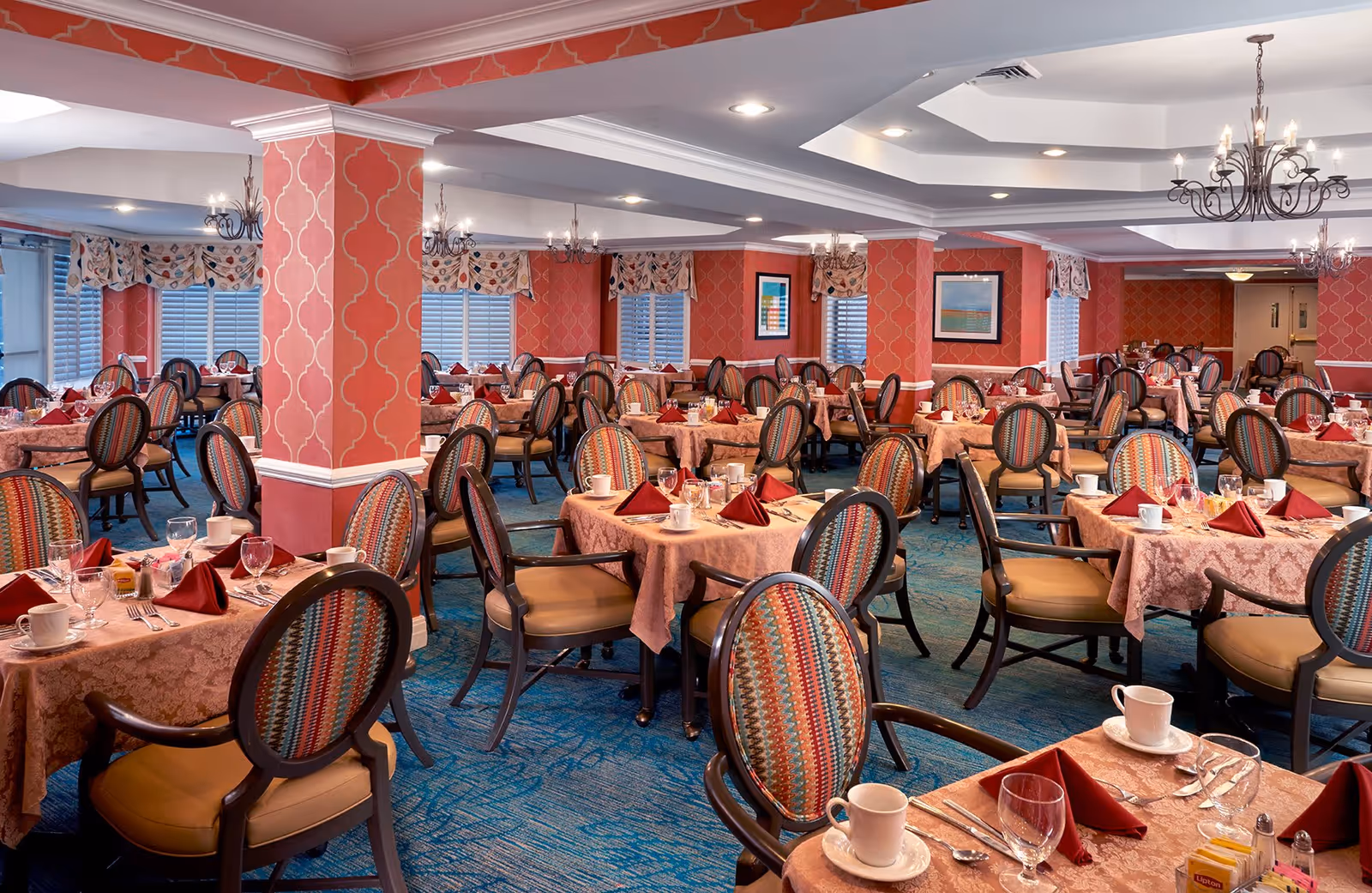 A spacious dining room with multiple round tables covered with pink tablecloths, each set with white cups, glasses, silverware, and folded red napkins. The chairs have patterned backs and cushioned seats. The walls are decorated with red wallpaper featuring a white geometric pattern, and there are several chandeliers hanging from the ceiling. Windows with patterned valances line the walls, and framed artwork is visible.