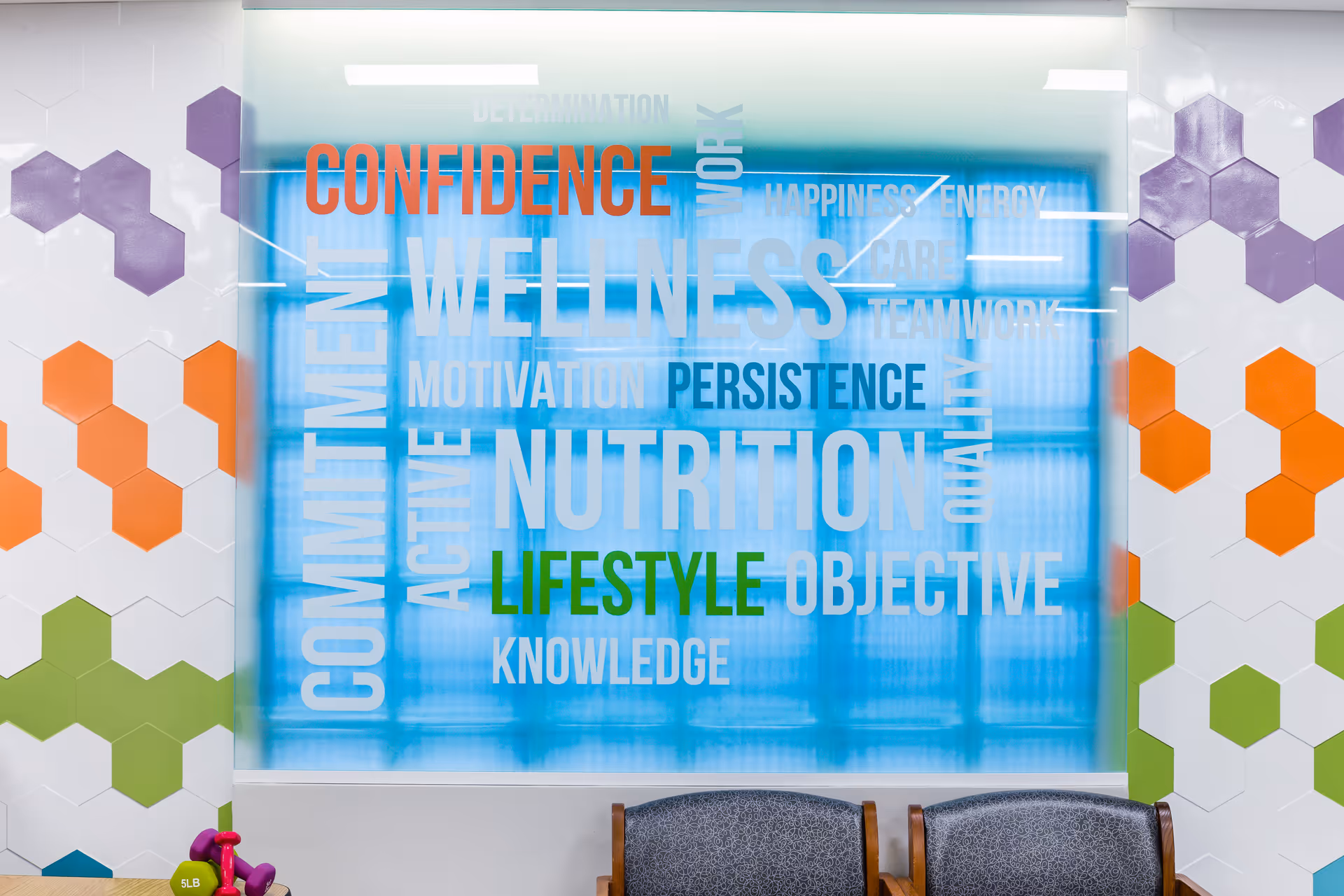Interior wall decorated with colorful hexagonal tiles in purple, orange, and green. A large frosted glass panel displays motivational words such as CONFIDENCE, WELLNESS, NUTRITION, LIFESTYLE, COMMITMENT, and others. Two chairs with patterned upholstery are partially visible at the bottom of the image.