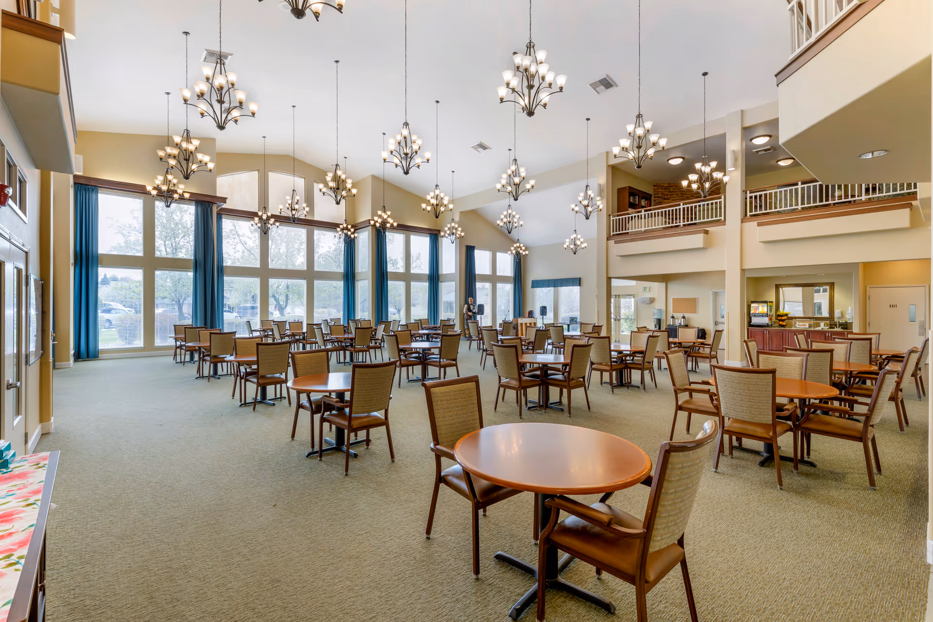 Spacious dining room with round tables and chairs, high vaulted ceilings, multiple chandeliers and large windows along one wall.