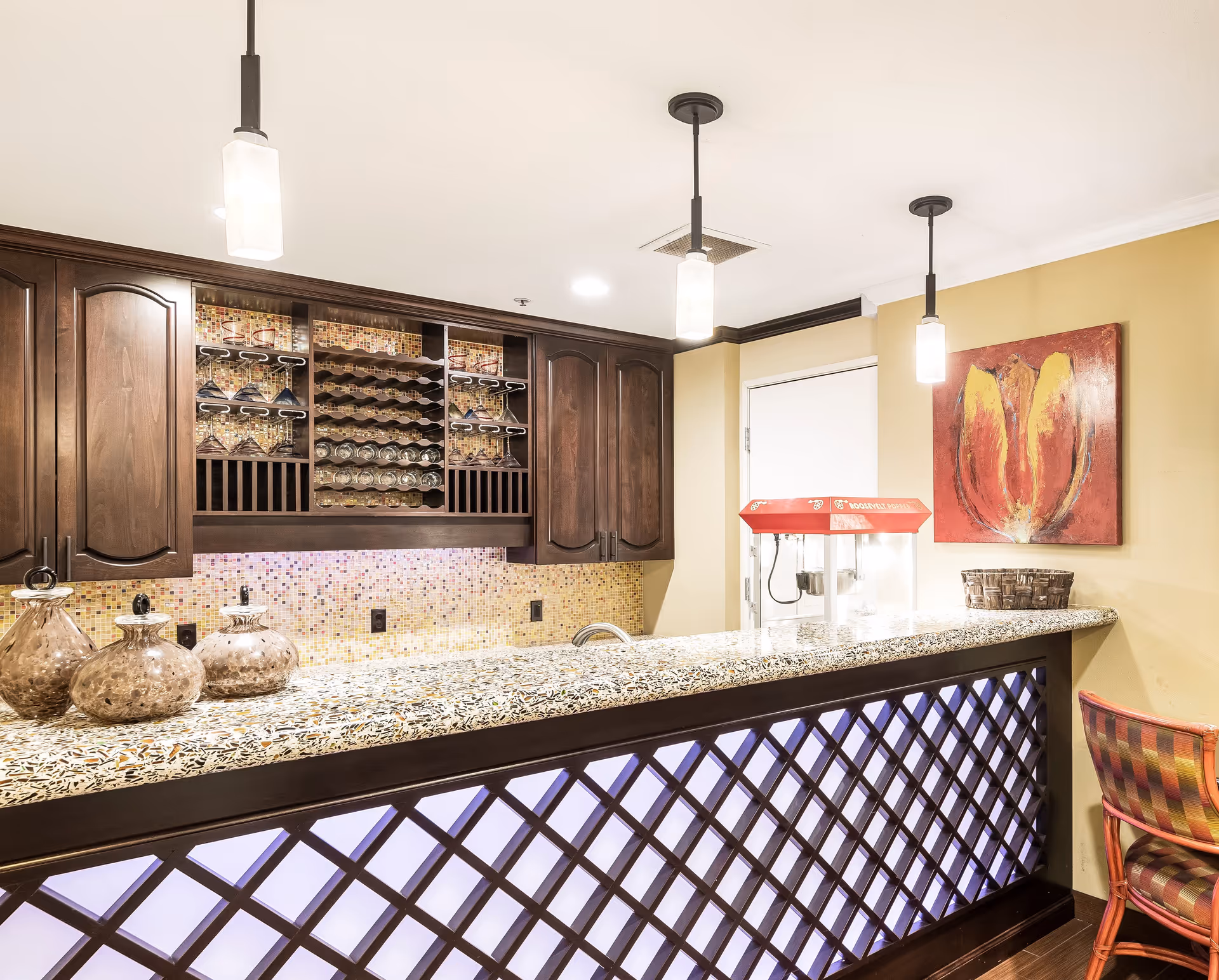 Interior view of a bar area with a granite countertop, dark wooden cabinets with wine racks and glass holders, three pendant lights hanging from the ceiling, a popcorn machine on the counter, a colorful abstract painting on the wall, and a chair with a patterned cushion.