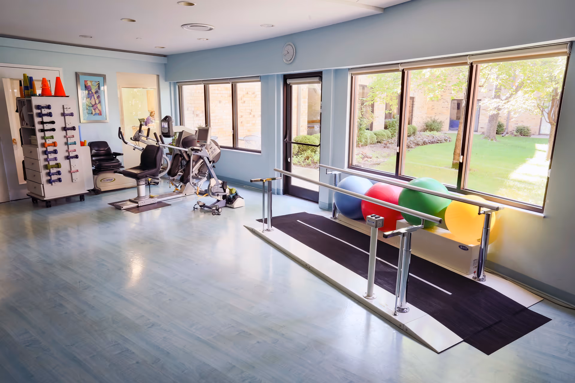 Physical therapy/exercise room with parallel bars, exercise bikes, a weight rack and colorful therapy balls by large windows overlooking a courtyard.