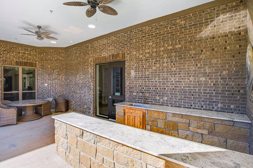 Covered outdoor patio with a stone bar countertop and sink, wicker seating, ceiling fans, and a brick wall backdrop.