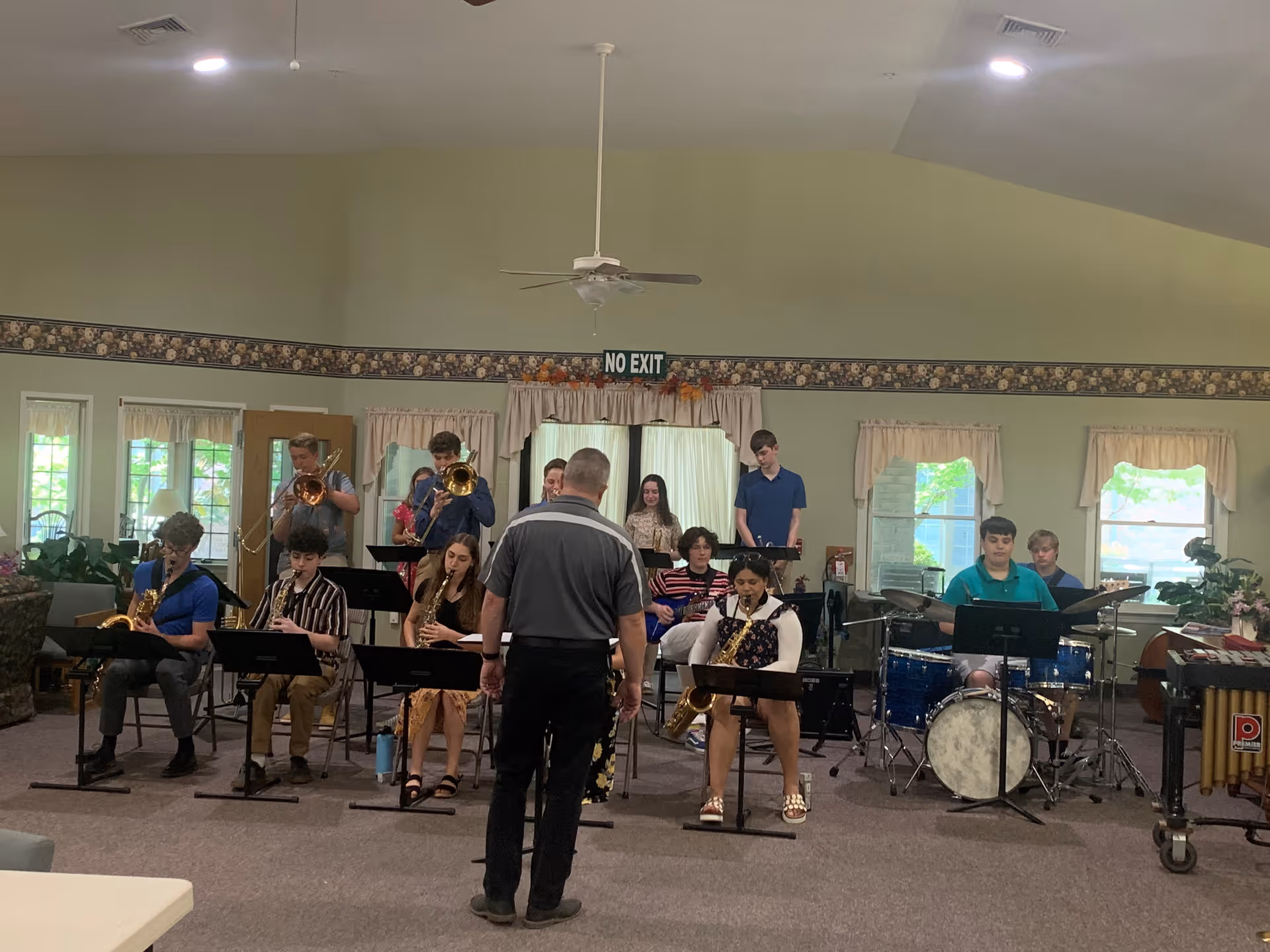 A group of young musicians playing various instruments including saxophones, trombones, drums, and guitars in a room with light green walls and floral wallpaper border. A conductor stands in front of them. The room has several windows with curtains and a ceiling fan.