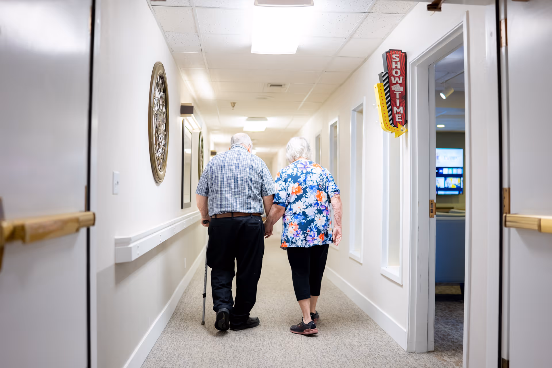 An elderly couple walking hand in hand down a well-lit hallway in a senior living facility. The man uses a cane and the woman wears a floral shirt. The hallway has white walls, handrails, and decorative wall art. A sign reading 'SHOWTIME' with an arrow is visible on the right side near an open door leading to a room with a television.