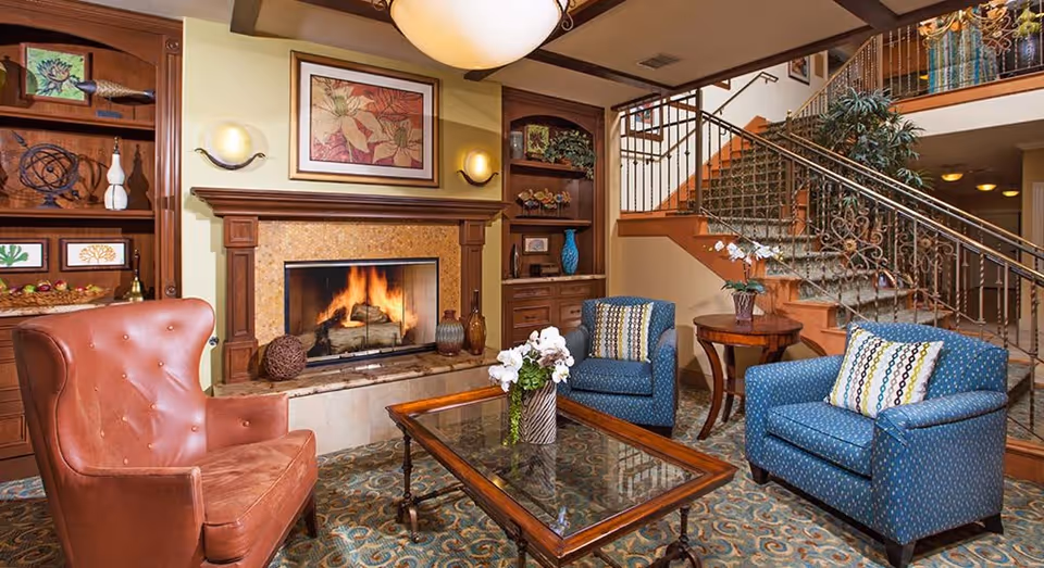 A cozy living room area with a lit fireplace surrounded by wooden shelves filled with decorative items. The room features a brown leather armchair, two blue upholstered armchairs with patterned pillows, a glass coffee table with a flower vase, and a wooden side table with another flower vase. A staircase with a decorative railing is visible in the background.