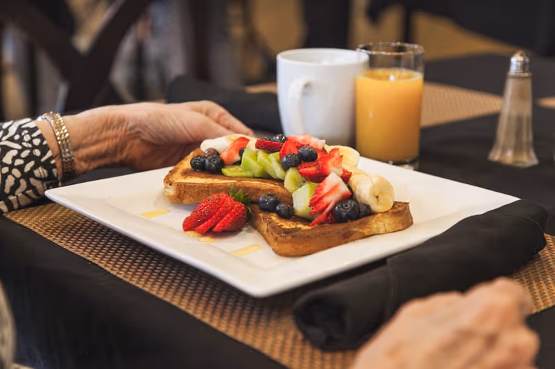 A close-up of a plate with two slices of toast topped with sliced bananas, strawberries, kiwi, and blueberries. The plate is on a table with a black napkin, a cup of coffee, a glass of orange juice, and a salt shaker. An elderly person's hands are visible near the plate.