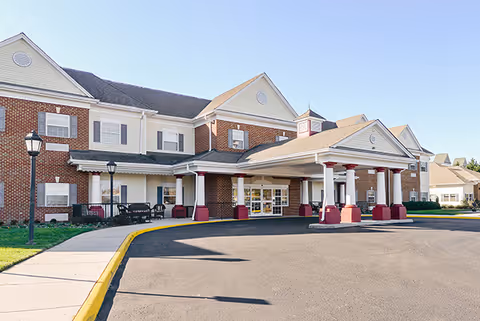 Front exterior view of a senior living facility building with a covered entrance supported by white columns with red bases, brick and beige siding, multiple windows, and a paved driveway with a yellow curb.