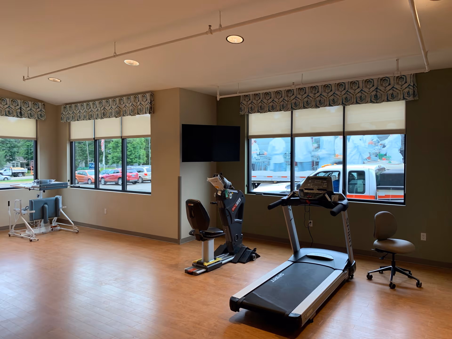 A fitness room with exercise equipment including a treadmill, a recumbent bike, and a standing exercise machine. The room has large windows with patterned valances, allowing natural light to enter. Outside the windows, parked cars and greenery are visible. There is a wall-mounted TV and a rolling office chair in the room.