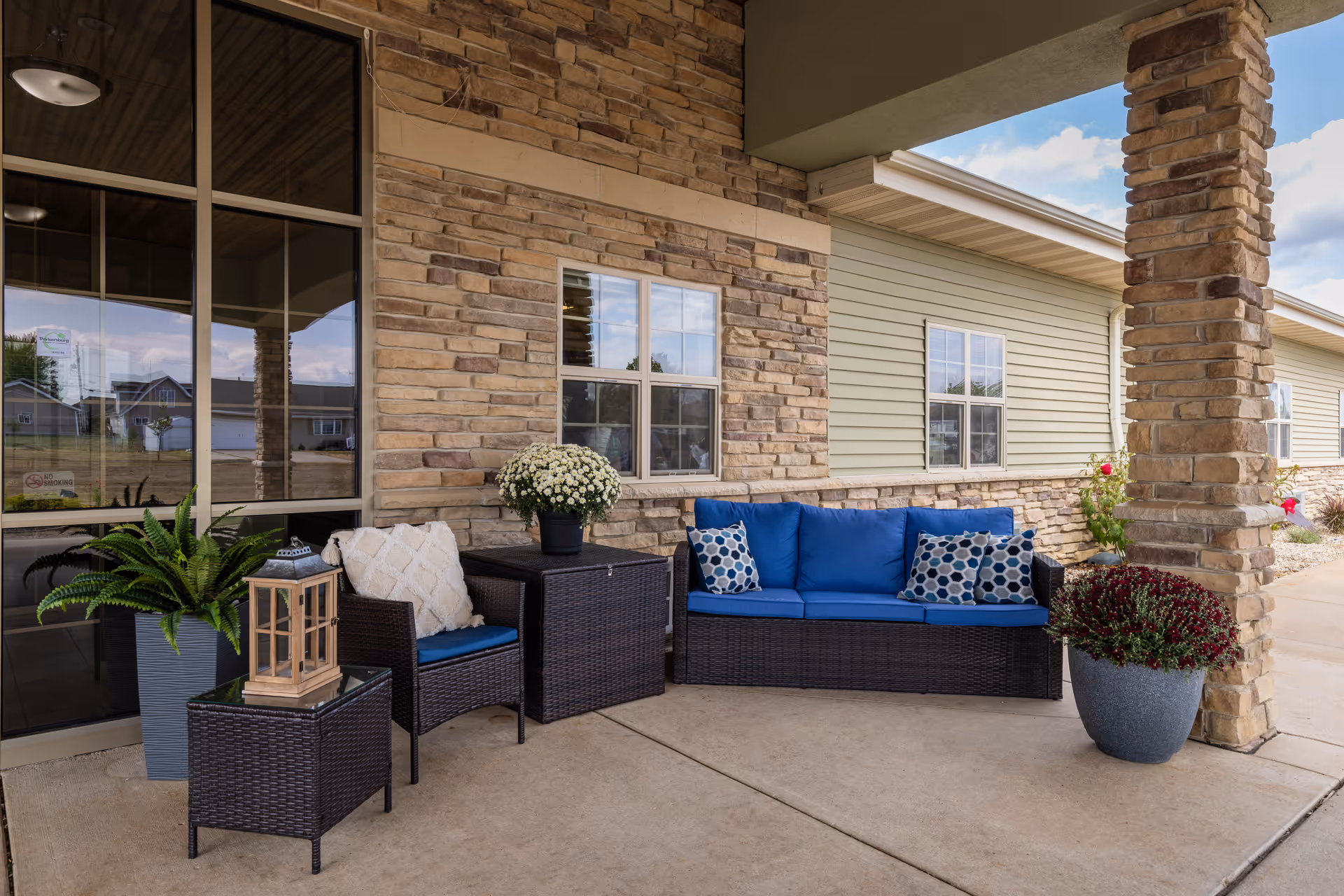 Outdoor seating area at Parker Place Retirement Community featuring a wicker sofa with blue cushions and patterned pillows, a matching chair with a white pillow, a small wicker table with a wooden lantern, and potted plants. The area is covered by a roof and has stone pillars and a stone wall with windows.