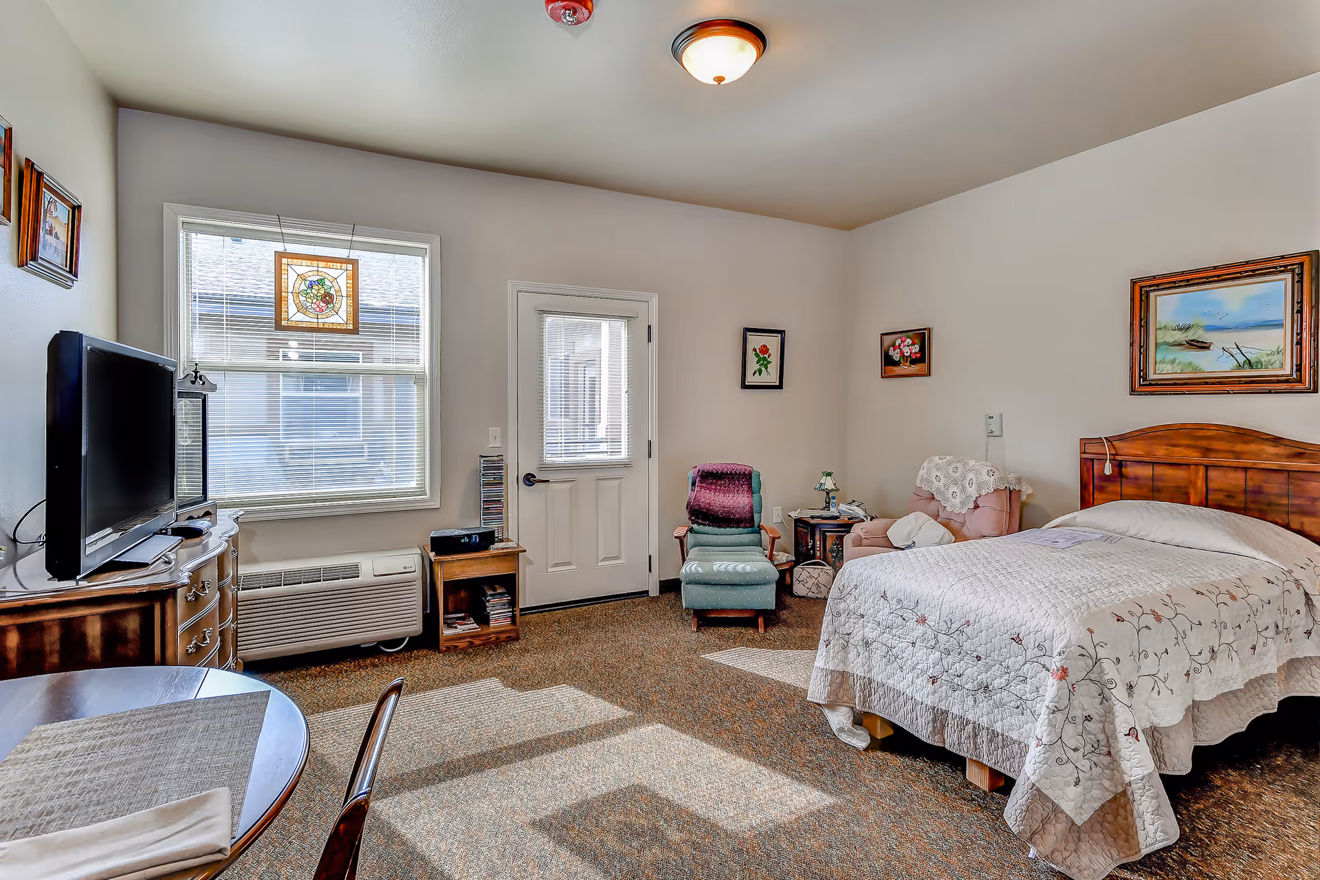 A cozy bedroom in an assisted living facility with a bed covered in a white quilt, a wooden headboard, a pink armchair with a white doily, a green recliner with a purple blanket, a small wooden side table with a lamp, a TV on a wooden dresser, a round table with a placemat and napkin, and a window with blinds and a stained glass decoration. The room has beige walls and carpeted floor.