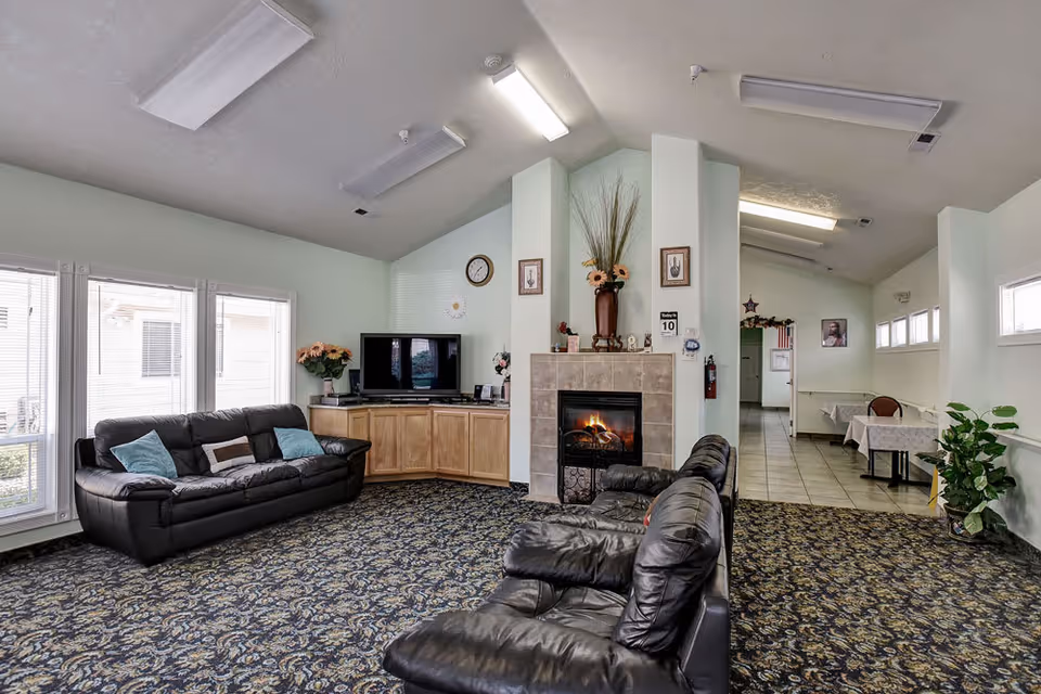 A cozy living room area with a patterned carpet, two black leather sofas with blue and brown cushions, a fireplace with a fire burning, a TV on a wooden cabinet, and large windows letting in natural light. The room has light-colored walls and ceiling with fluorescent lights, and there is a hallway leading to another area with tables covered in white tablecloths.
