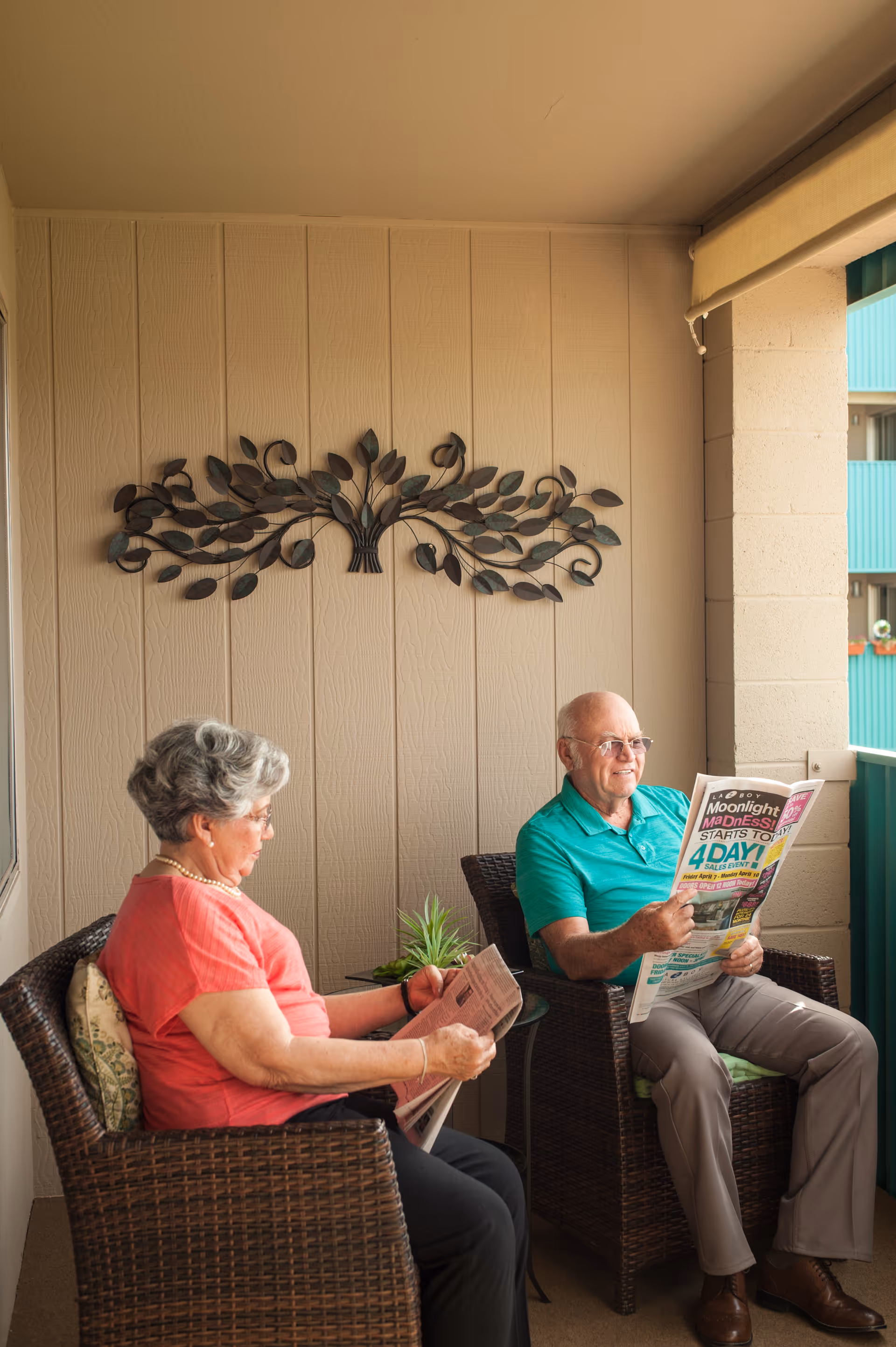 An elderly man and woman sitting on wicker chairs on a covered balcony or patio, both reading newspapers. The wall behind them is beige with a decorative metal wall art featuring leaves. There is a small glass table with a plant between them.