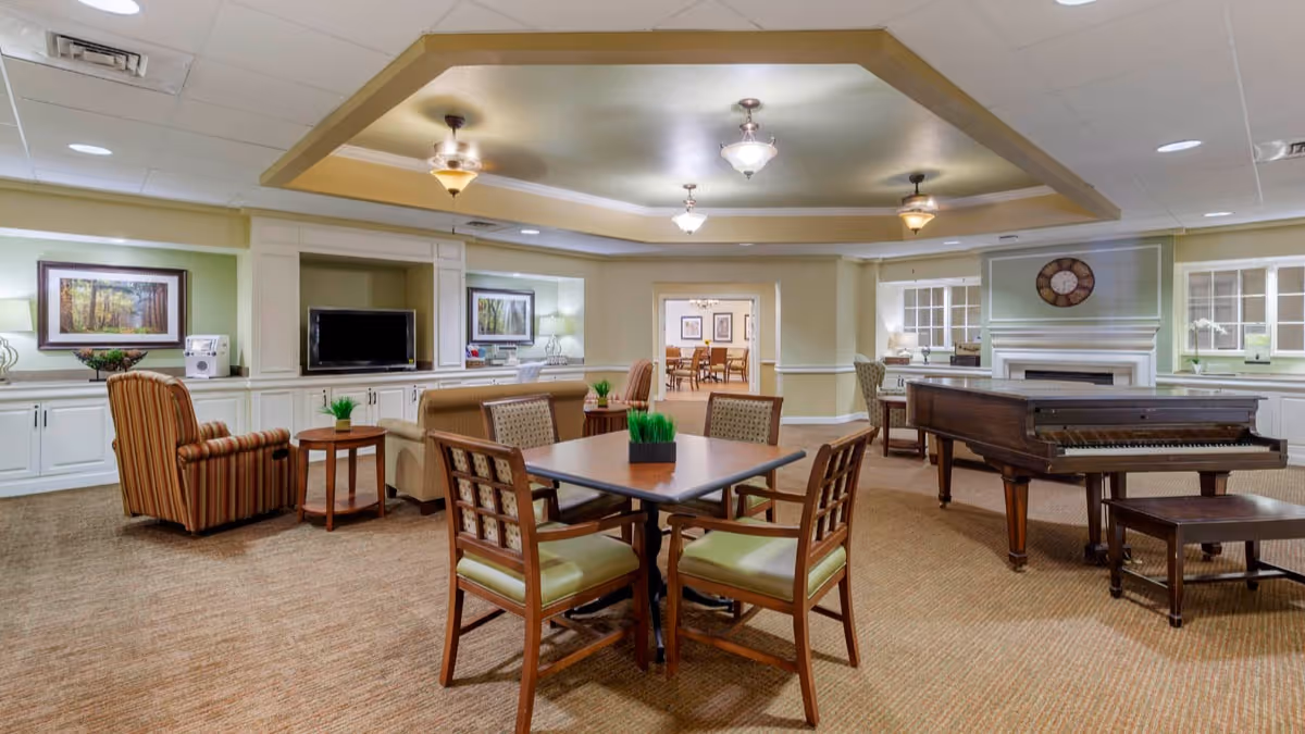 Bright communal living room with tables and chairs, upholstered seating, a grand piano, TV, and fireplace.