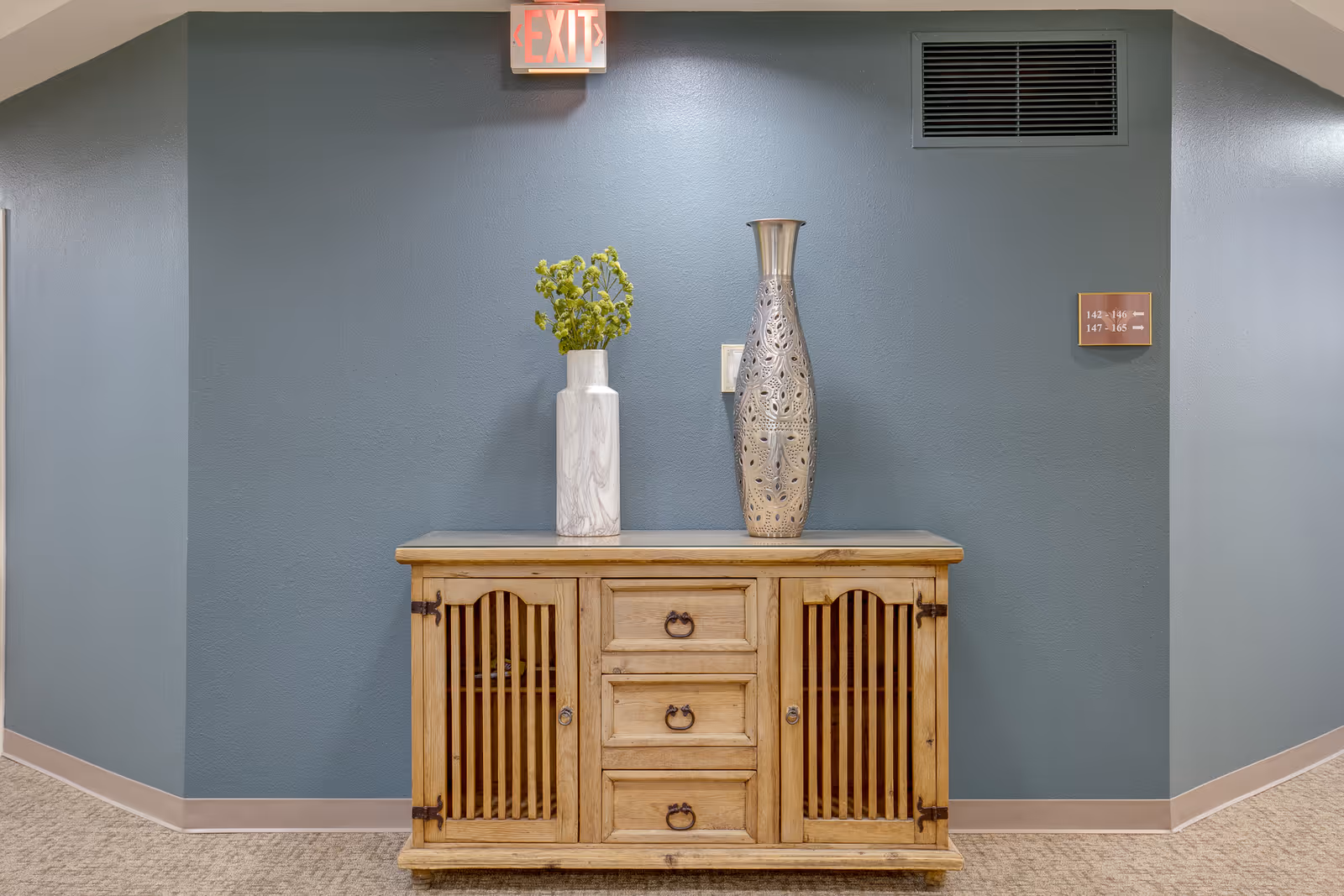 A wooden cabinet with three drawers and two slatted doors stands against a blue-gray wall. On top of the cabinet are two decorative vases, one white with green foliage and the other tall and metallic with intricate cut-out patterns. Above the cabinet is an illuminated exit sign, and to the right on the wall is a small sign with room numbers and arrows. The floor is carpeted in a neutral tone.