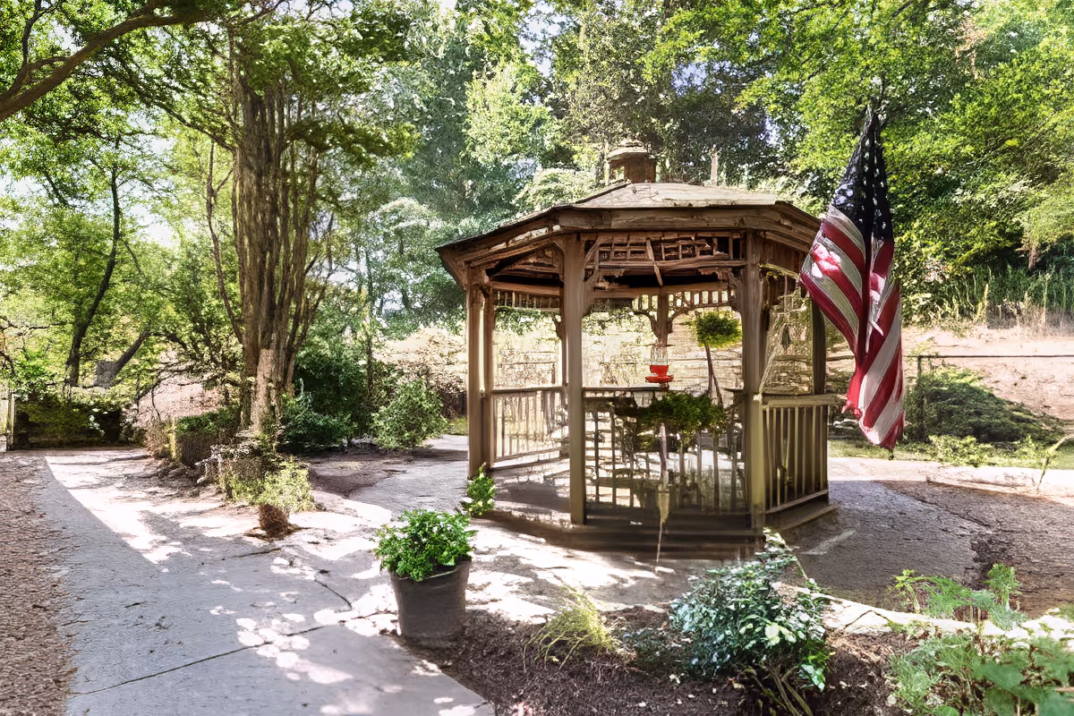 Wooden gazebo in a landscaped garden with an American flag, surrounding trees, and paved walkways.