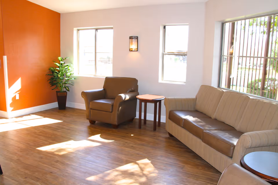 A bright living room area with wooden flooring, an orange accent wall, a potted plant, a brown leather armchair, a beige sofa with brown cushions, and two small round wooden tables. Sunlight streams in through three windows with bars on the outside.
