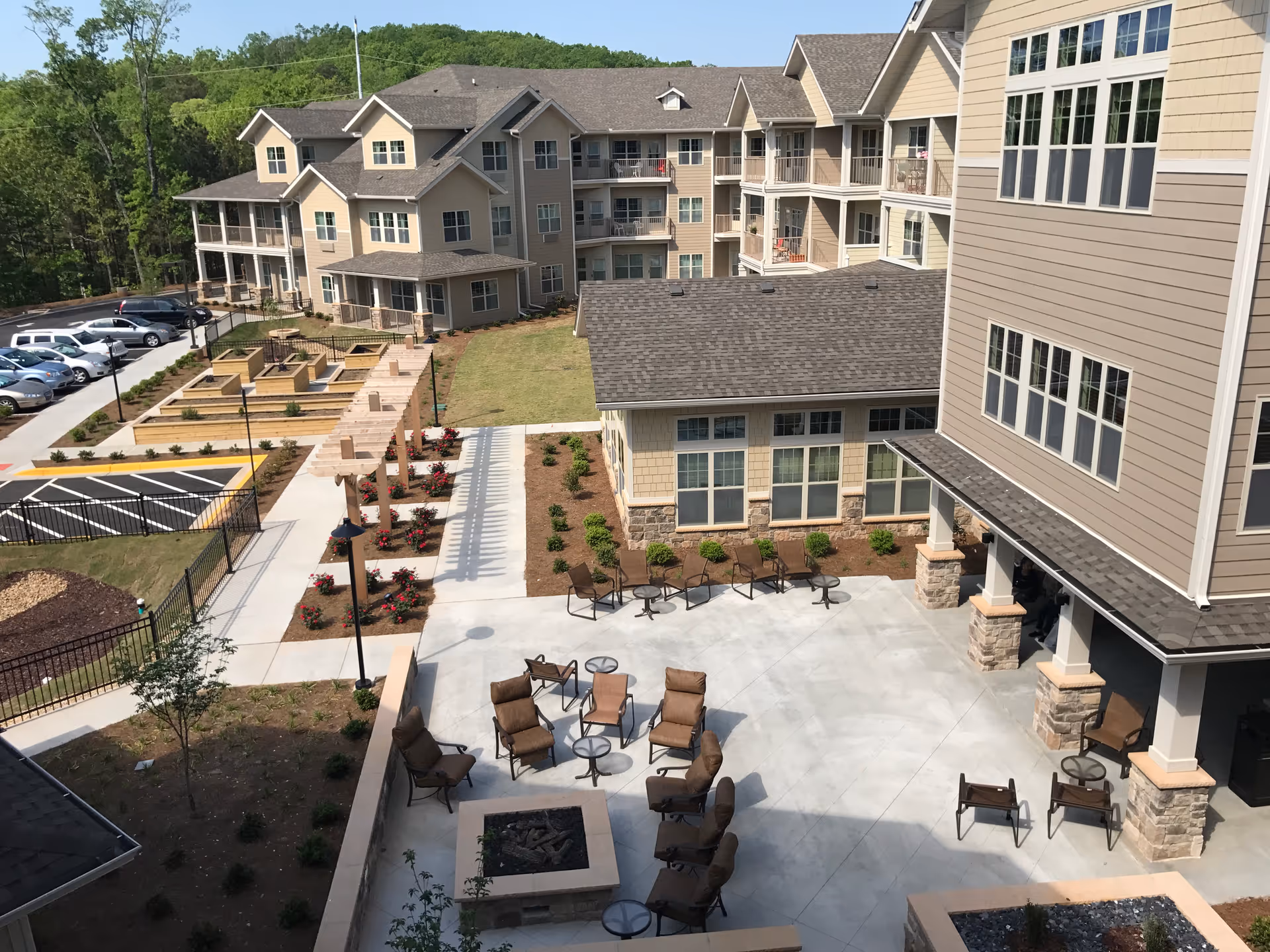 Outdoor courtyard area of a senior living facility with multiple beige buildings surrounding a concrete patio. The patio has several brown cushioned chairs and small round tables arranged around a fire pit. There is a pergola with rose bushes along a walkway, a parking lot with cars, and green trees in the background.