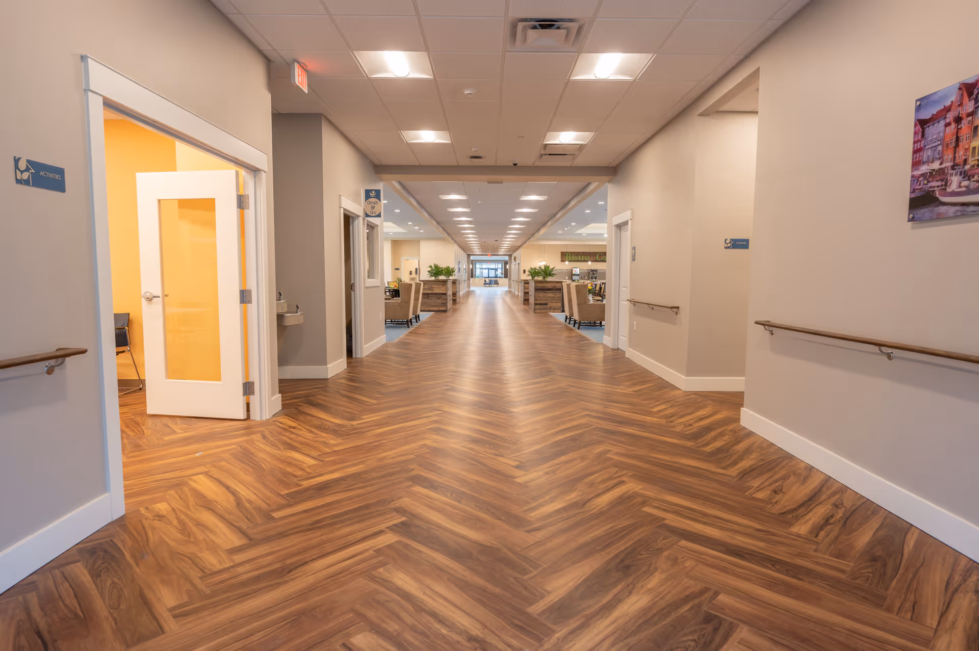 A wide hallway in a senior living facility with wood-patterned flooring and beige walls. On the left, there is an open door leading to a room labeled 'Activities' with yellow walls and chairs inside. On the right, there is a door labeled 'Storage' and a colorful framed picture on the wall. The hallway extends forward with seating areas and plants visible in the distance, and ceiling lights illuminating the space.