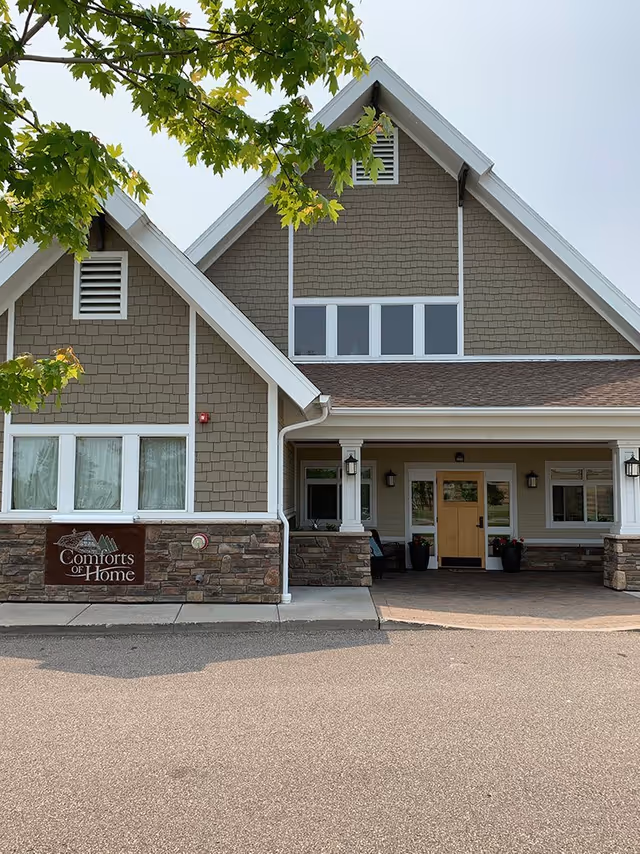 Exterior front view of a senior living facility building with a peaked roof, beige siding, stone accents, and a covered entrance. There is a sign on the left side of the building that reads 'Comforts of Home'. Green tree branches partially frame the top of the image.