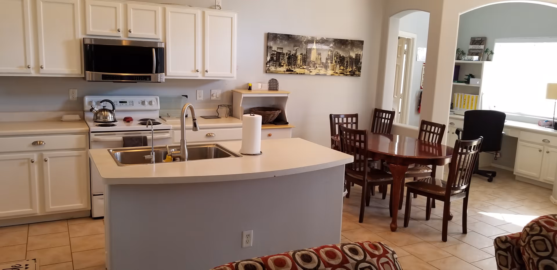 Interior view of a kitchen and dining area in an assisted living facility. The kitchen features white cabinets, a stove with a kettle, a microwave, and a sink on a central island with a paper towel holder. Adjacent to the kitchen is a wooden dining table with six chairs. In the background, there is a desk with a black office chair and shelves with books and decor. The floor is tiled, and the walls are light-colored with a cityscape painting hanging above a small cabinet.