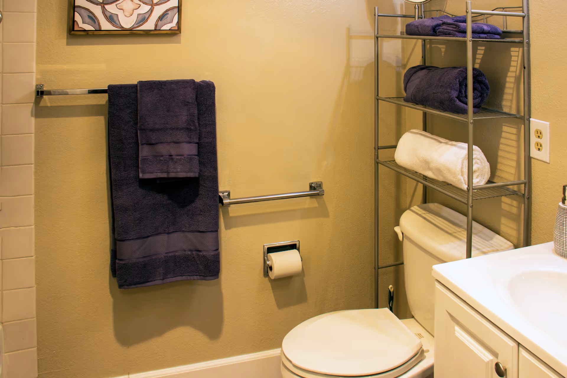 Modern bathroom with toilet, sink, towel rack, and metal shelving holding folded towels.