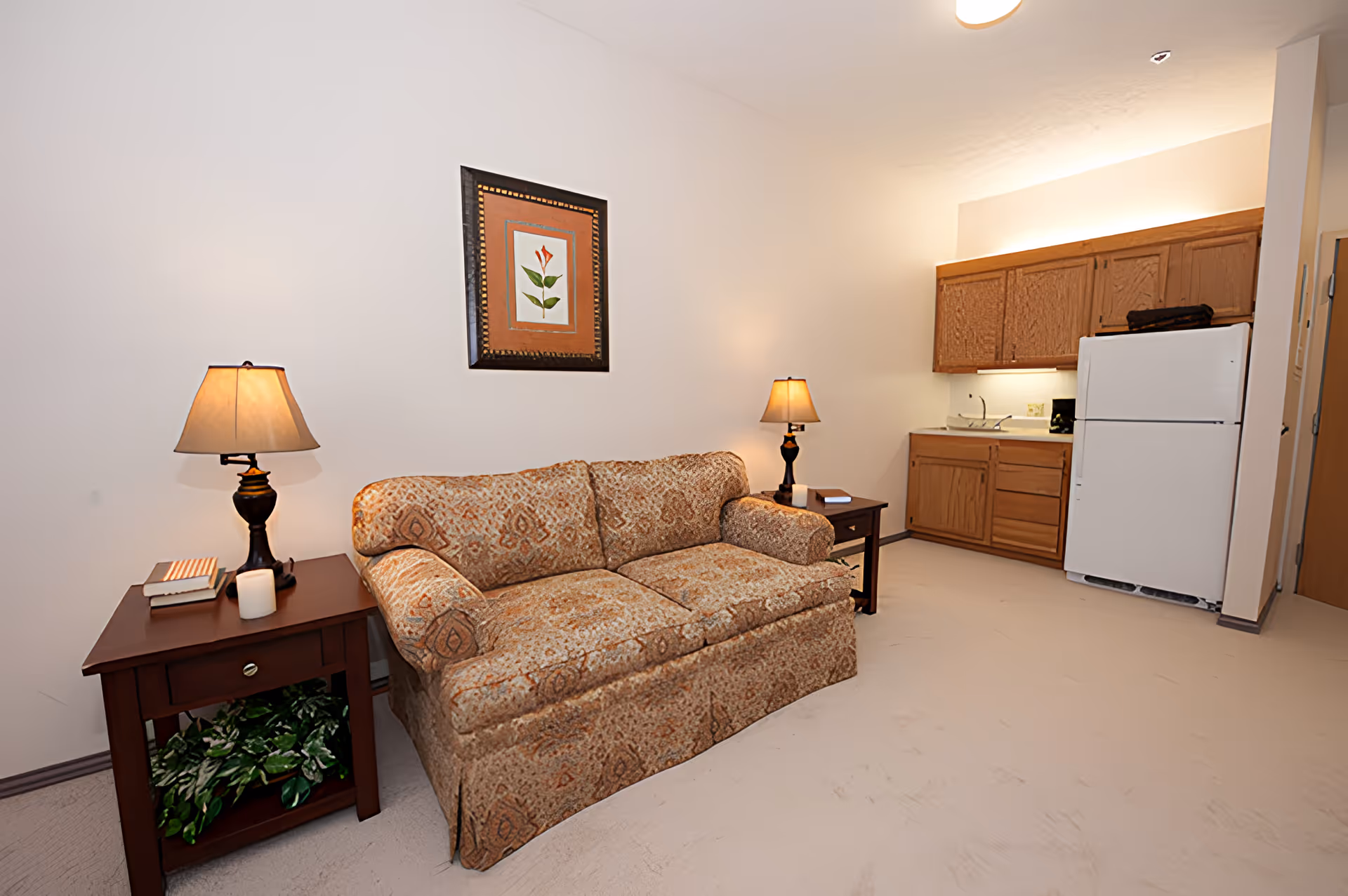 Small living area with a patterned sofa, side tables and lamps, and a kitchenette with refrigerator and cabinets.