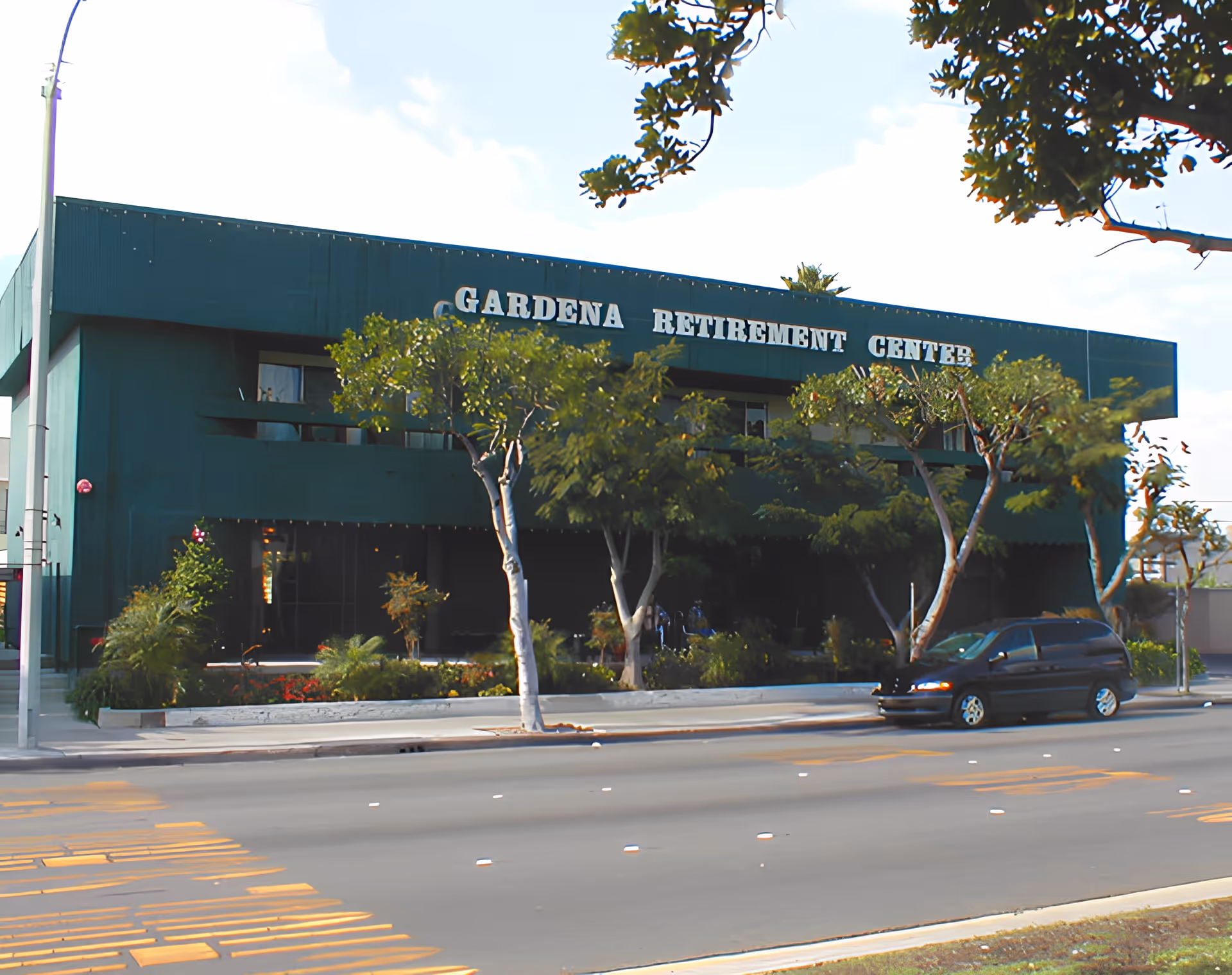 Exterior front view of a two-story green building with the sign 'Gardena Retirement Center' on the upper facade. There are several trees and plants in front of the building, and a black minivan is parked on the street nearby.