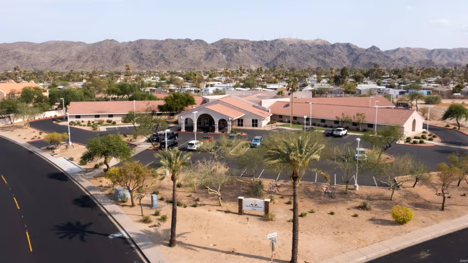 Aerial view of South Mountain Post Acute facility showing a single-story building with a red-tiled roof, surrounded by a parking lot with several cars. The facility is located in a desert landscape with palm trees and sparse vegetation, with mountains visible in the background under a clear sky.