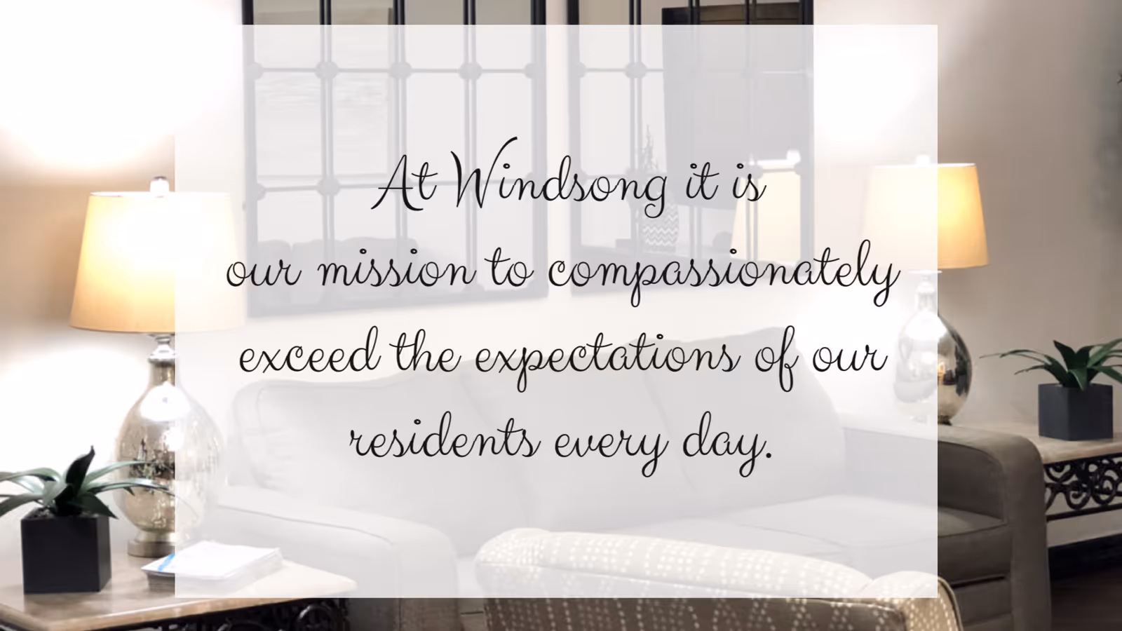 A cozy living room with a white sofa, two table lamps with beige shades on side tables, a small green plant, and a decorative mirror on the wall. Overlaid text reads: 'At Windsong it is our mission to compassionately exceed the expectations of our residents every day.'