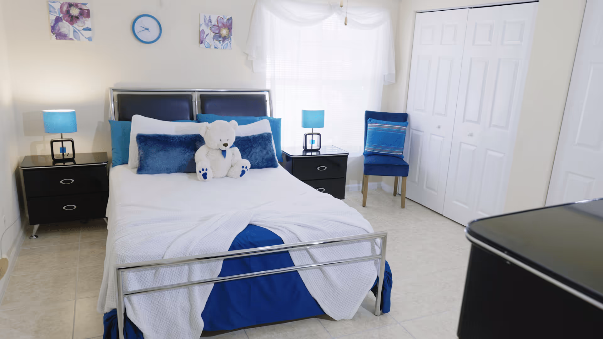 A neatly made bed with white and blue bedding and a white teddy bear sitting in the center. The room has two black nightstands with blue lamps on each side of the bed. There is a blue chair with a decorative pillow next to a window with white curtains. The walls are light-colored with two floral paintings and a blue clock above the bed. The floor is tiled and there are white double closet doors on the right side.