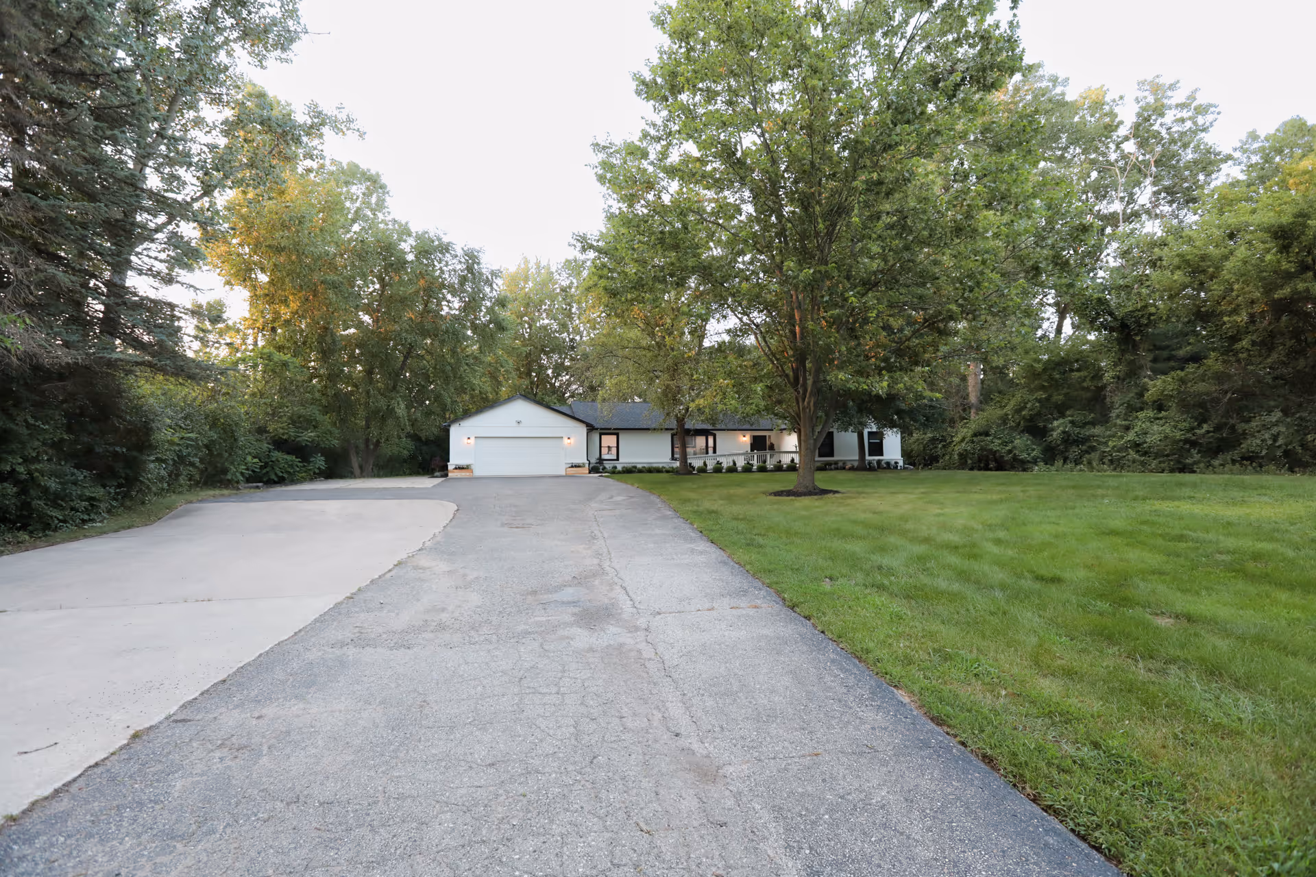A single-story white building with a two-car garage at the end of a long driveway, surrounded by green grass and tall trees under a clear sky.