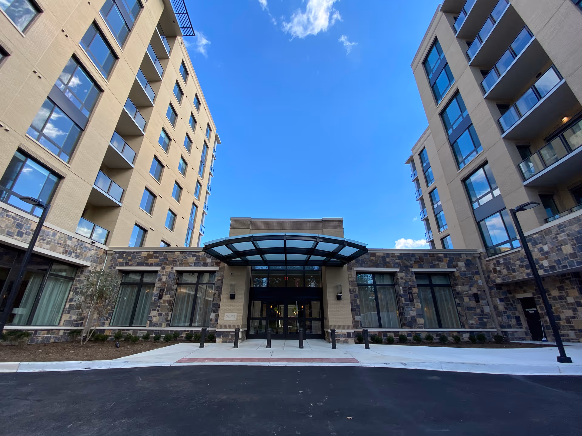 Front entrance of a multi-story residential building with a glass canopy between two wings under a bright blue sky.