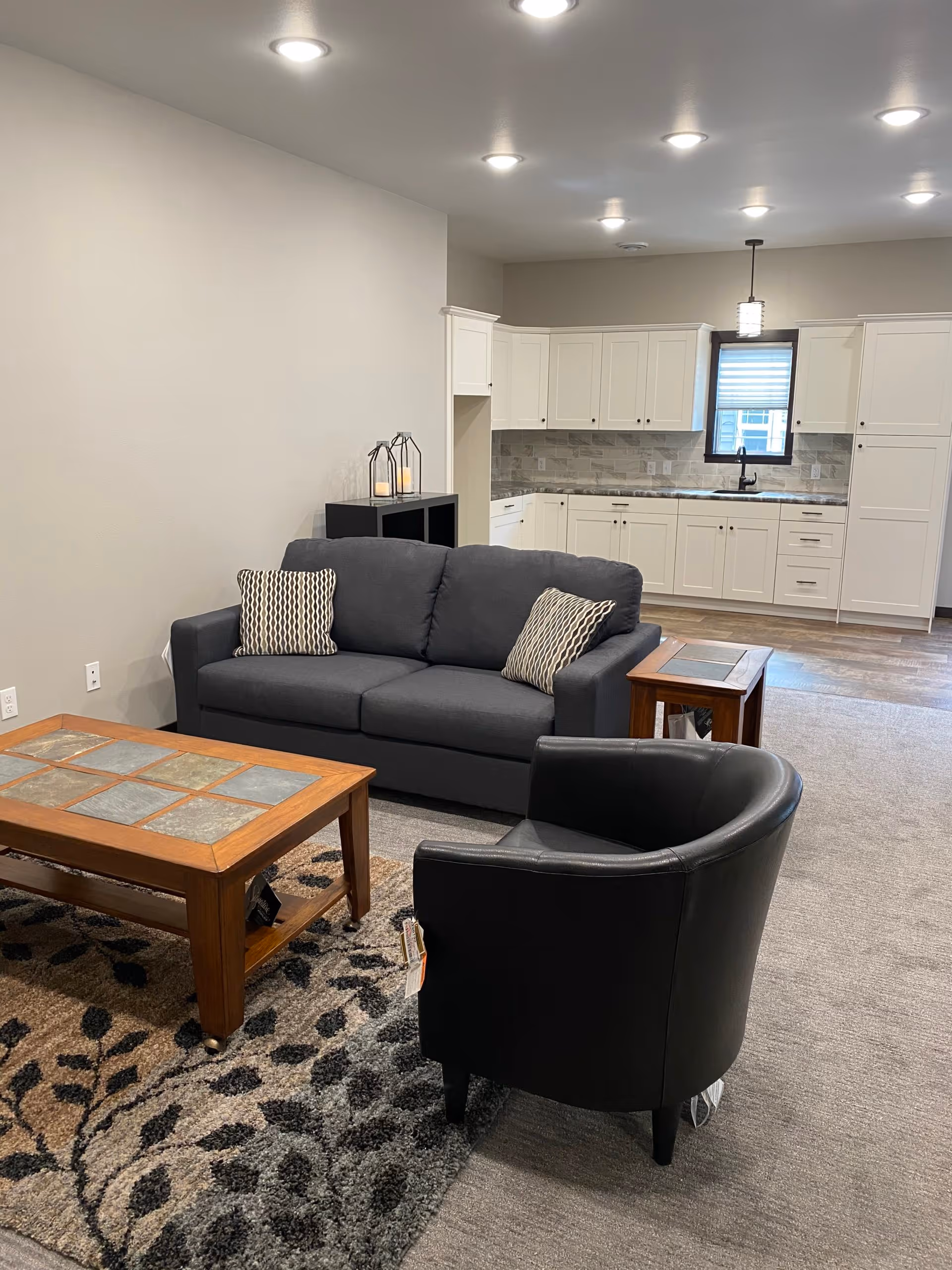 A furnished living area with a gray sofa, black armchair, wooden coffee table and rug, with a white kitchen visible in the background.