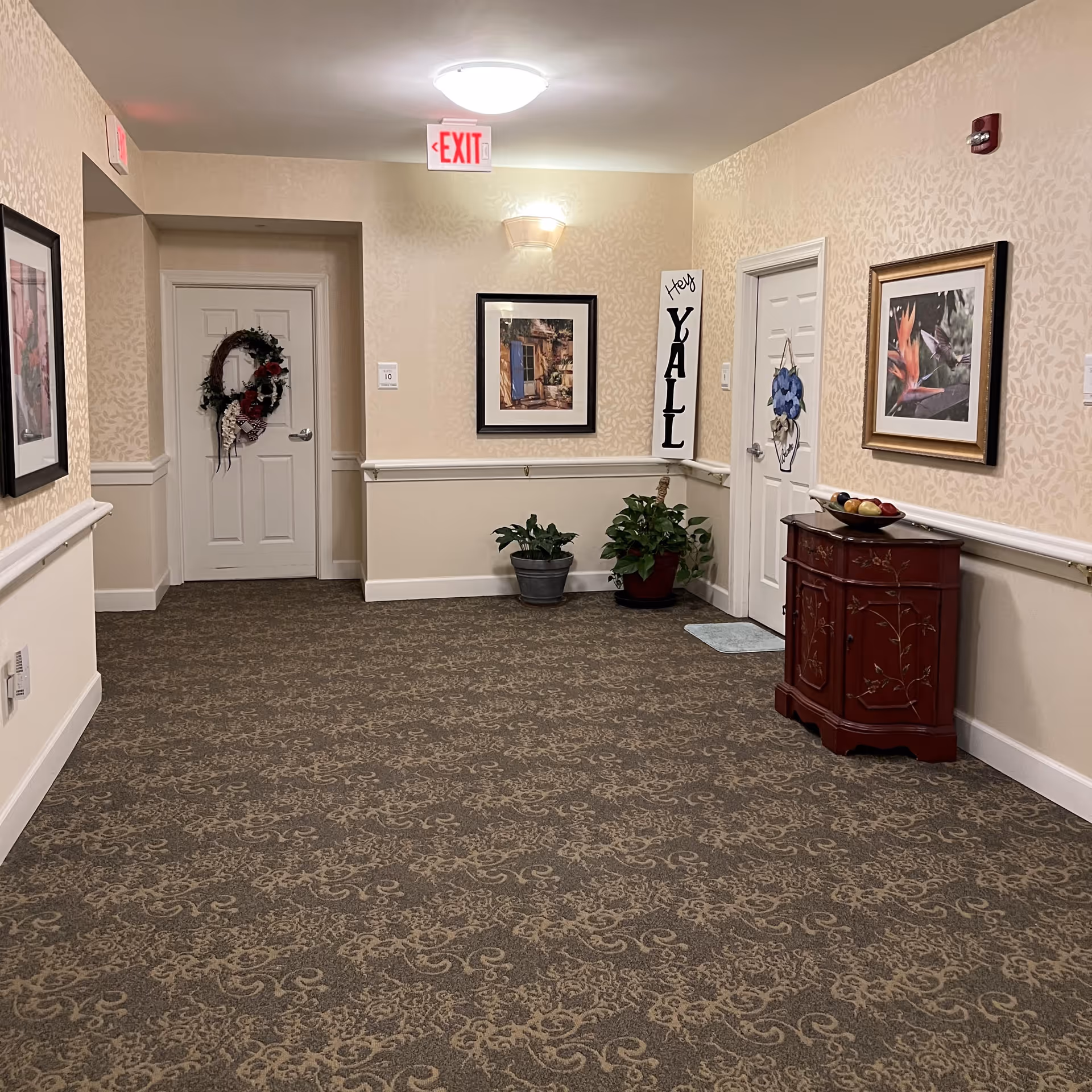 A carpeted hallway in an assisted living facility with beige patterned wallpaper and white trim. There are two closed white doors, one decorated with a wreath and the other with a hanging flower decoration. The hallway features framed artwork on the walls, two potted plants, a small wooden cabinet with a bowl of fruit on top, and a vertical sign that says 'Hey YALL'. An illuminated exit sign is visible on the ceiling.
