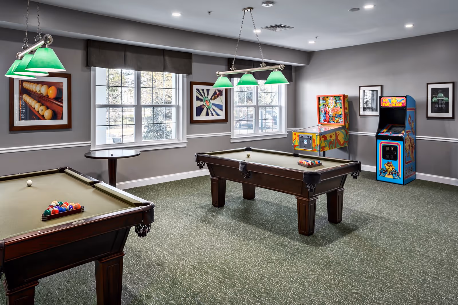 A recreational room with two pool tables, a pinball machine, and a Ms. Pac-Man arcade game. The room has green carpet, gray walls with white trim, framed pictures on the walls, and large windows letting in natural light. Green hanging lights illuminate the pool tables.