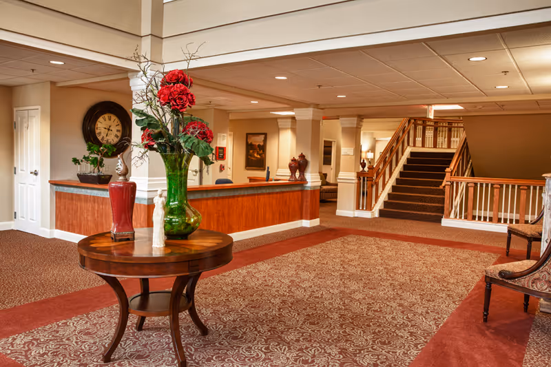 A senior living facility reception lobby with a round table holding a large vase of red flowers, a front desk, and a staircase in the background.